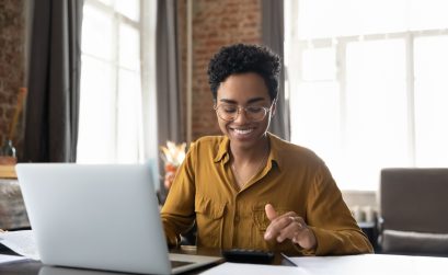 Happy young Afro American entrepreneur woman in glasses counting profit, on calculator at laptop computer, analyzing benefits, enjoying financial success, job high result, smiling
