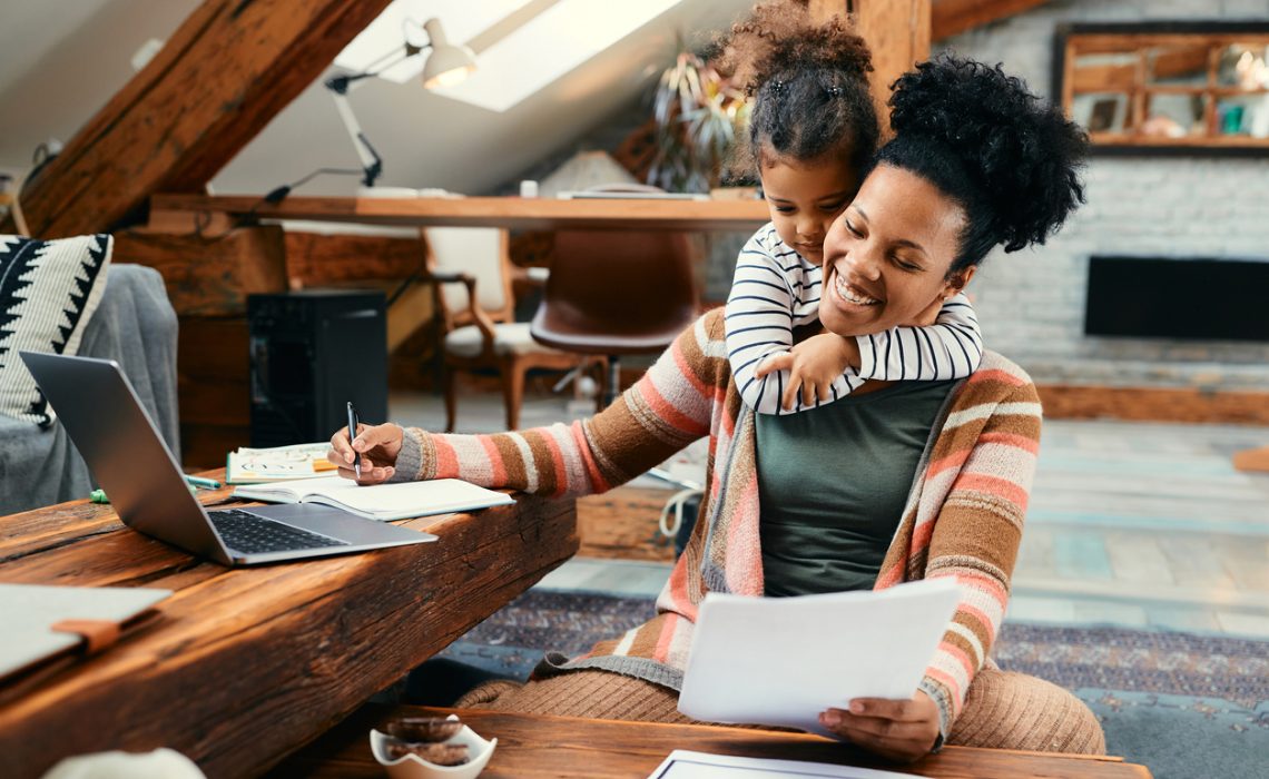 Happy African American mother using laptop and working on paperwork while her daughter is embracing her at home.